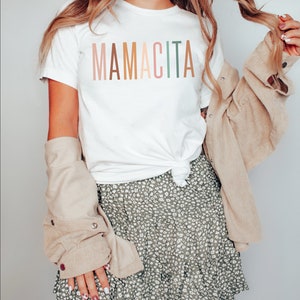May include: White t-shirt with the text "MAMACITA" in rainbow colors. The shirt is worn by a person with a green floral skirt and a beige jacket.