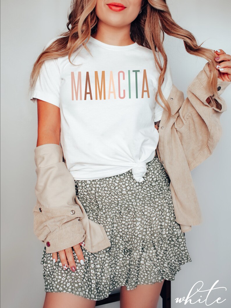 May include: White t-shirt with the text "MAMACITA" in rainbow colors. The shirt is worn by a person with a green floral skirt and a beige jacket.