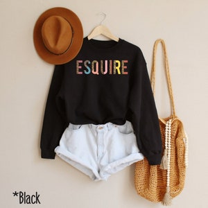Peut inclure: Sweat-shirt noir avec le mot "ESQUIRE" imprim&eacute; en couleurs arc-en-ciel avec un motif l&eacute;opard. Le sweat-shirt est port&eacute; avec un short en jean blanc et un sac en crochet marron.