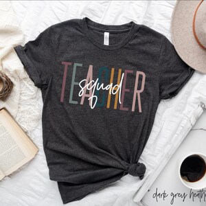 Peut inclure: Un t-shirt gris foncé chiné avec le texte "Teacher Squad" en rayures verticales colorées. Le texte est en blanc.
