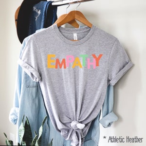 May include: Athletic heather gray t-shirt with the word "EMPATHY" in colorful block letters. The shirt is hanging on a wooden hanger with other clothing items in the background. The text "* Athletic Heather" is in the bottom right corner.