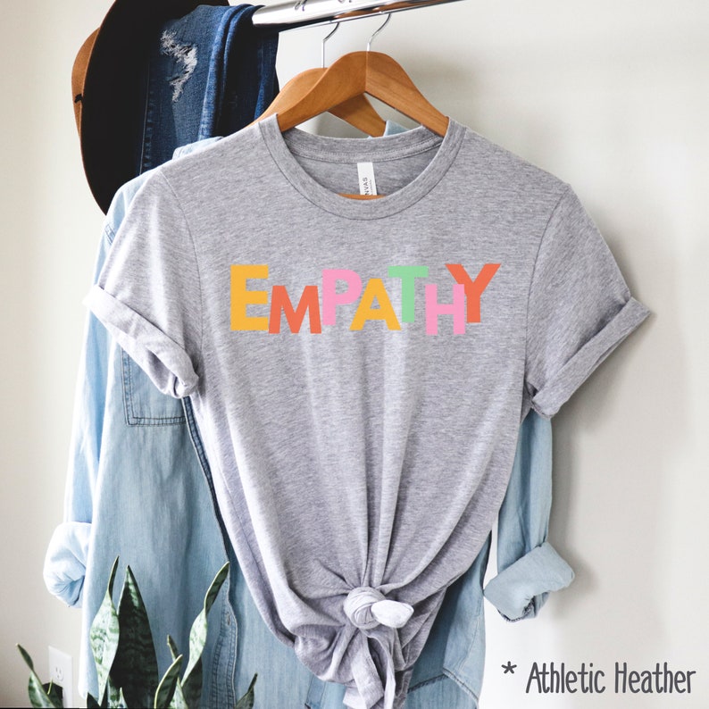 May include: Athletic heather gray t-shirt with the word "EMPATHY" in colorful block letters. The shirt is hanging on a wooden hanger with other clothing items in the background. The text "* Athletic Heather" is in the bottom right corner.