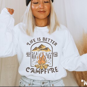 K&ouml;nnte beinhalten: Wei&szlig;er Pullover mit einem Grafikdruck einer Feuerstellenszene und dem Text "Life is better by the campfire".