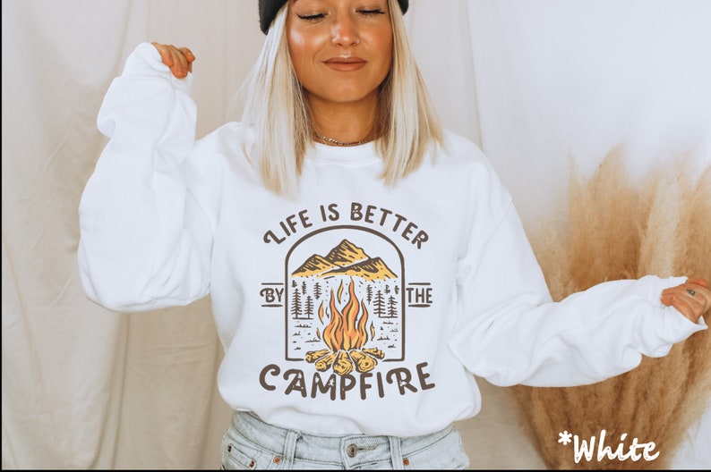 K&ouml;nnte beinhalten: Wei&szlig;er Pullover mit einem Grafikdruck einer Feuerstellenszene und dem Text "Life is better by the campfire".