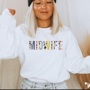 Pode incluir: Camisola branca com a palavra "MIDWIFE" impressa em letras coloridas com um padr&atilde;o de leopardo.