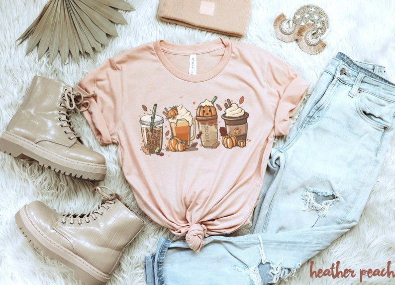May include: A pink t-shirt with a graphic of four fall-themed drinks, including a pumpkin spice latte, a caramel apple cider, and a peppermint mocha. The graphic also includes fall leaves and pumpkins.