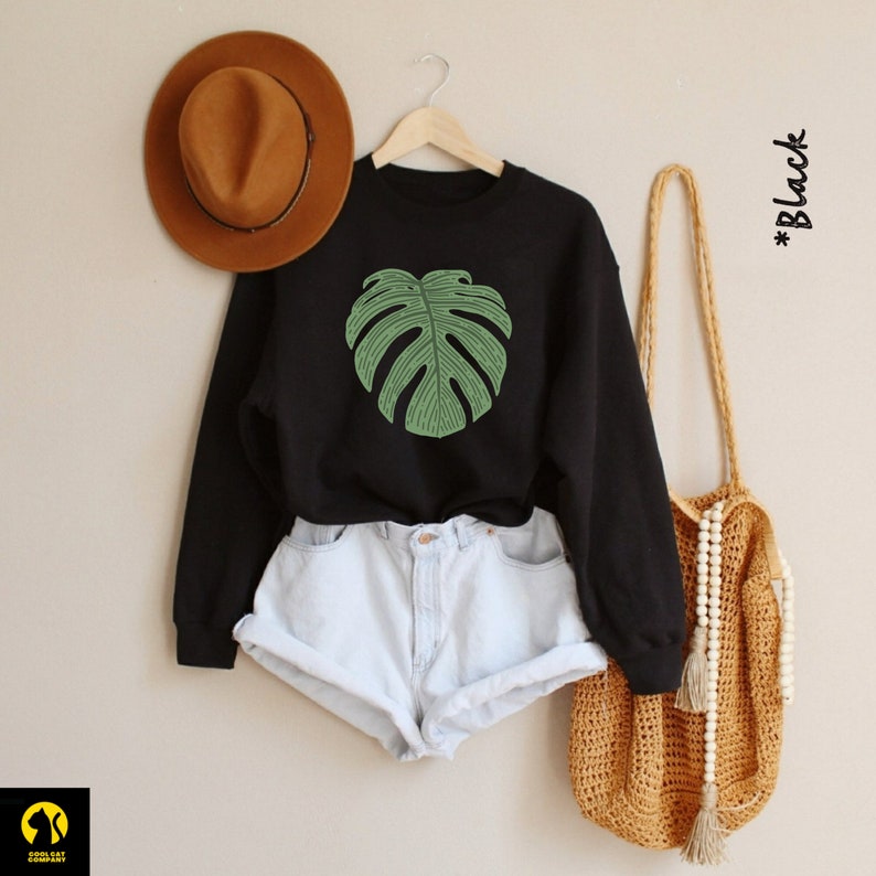 Peut inclure: Sweat-shirt noir avec un motif de feuille de monstera verte. Le sweat-shirt est port&eacute; avec un short en jean bleu clair et un chapeau marron. Le mod&egrave;le porte &eacute;galement un sac en crochet avec des perles en bois.