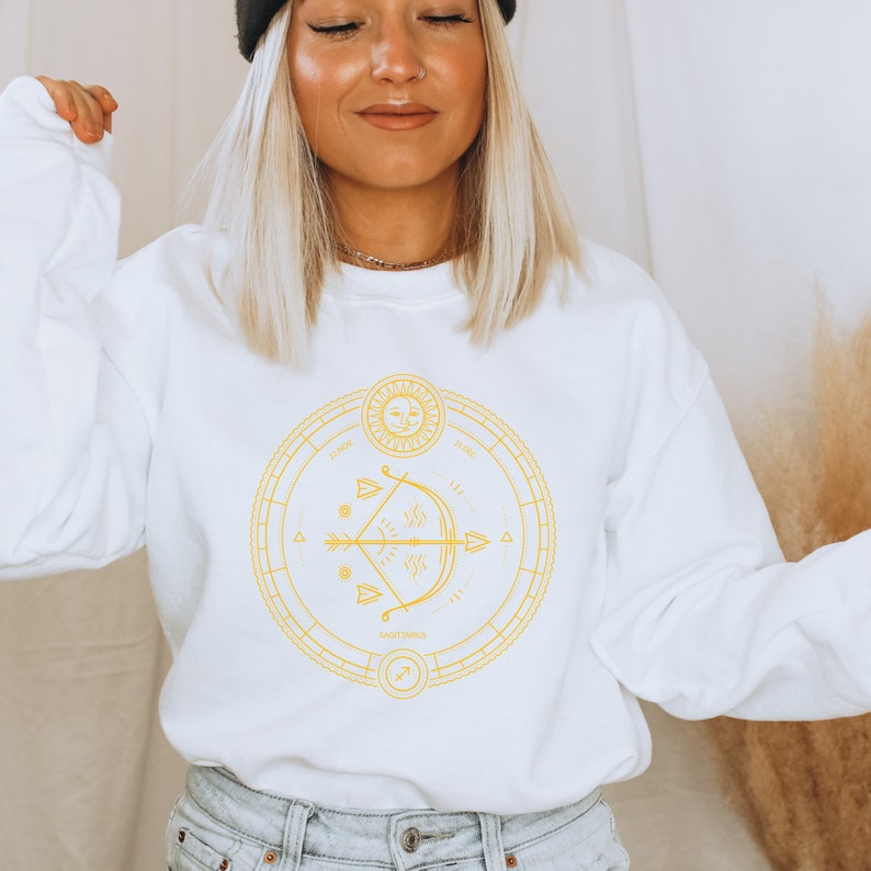 May include: White sweatshirt with a gold graphic of a zodiac symbol, an arrow and bow, and the words "Sagittarius" and "22 Nov - 21 Dec".