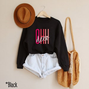 May include: Black sweatshirt with the text "OHH deer" in pink and red lettering. The sweatshirt is worn by a model wearing denim shorts.