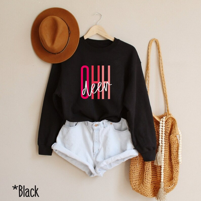 May include: Black sweatshirt with the text "OHH deer" in pink and red lettering. The sweatshirt is worn by a model wearing denim shorts.