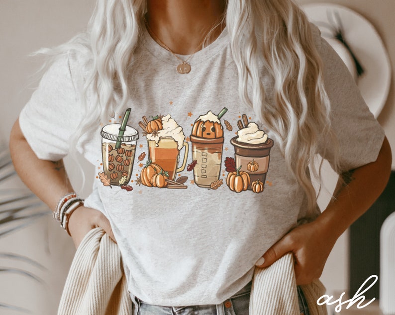 May include: A gray t-shirt with a graphic of four fall-themed drinks. The drinks are a pumpkin spice latte, a caramel macchiato, a pumpkin iced coffee, and a hot chocolate. Each drink is topped with whipped cream and a pumpkin. The graphic is surrounded by fall leaves and stars.