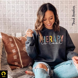 May include: A dark heather grey sweatshirt with the text "THERAPY is cool" in colorful letters. The letters are outlined in a leopard print pattern.