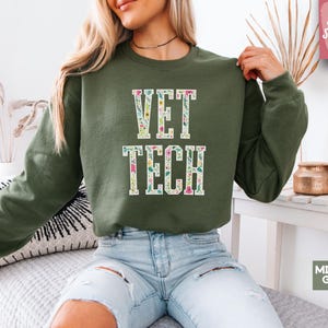 Puede incluir: Una sudadera verde con el texto "VET TECH" en letras blancas con contorno rosa. Las letras están decoradas con un patrón floral. La sudadera la lleva puesta una persona con el pelo largo y rubio.