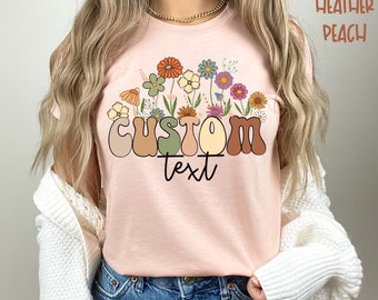Könnte beinhalten: Ein T-Shirt in der Farbe Heidepfirsich mit einer Retro-Grafik des Wortes "CUSTOM" in bunten Buchstaben, umgeben von Blumen und Blättern. Der Text "text" steht unter dem Wort "CUSTOM".
