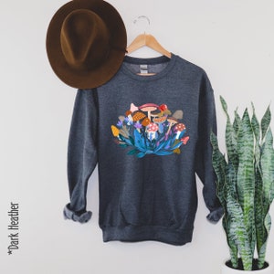 May include: Dark heather grey sweatshirt featuring a colourful mushroom graphic. The mushrooms are surrounded by green leaves and flowers.