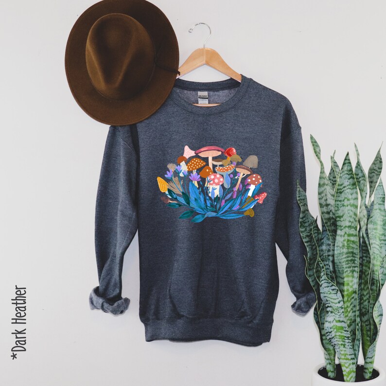 May include: Dark heather grey sweatshirt featuring a colourful mushroom graphic. The mushrooms are surrounded by green leaves and flowers.