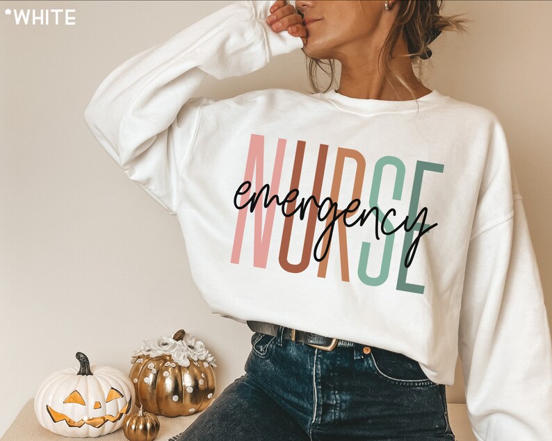 Op de afbeelding: Witte trui met de tekst "NURSE emergency" in kleurrijke verticale letters. De letters zijn roze, bruin, groen en wit.