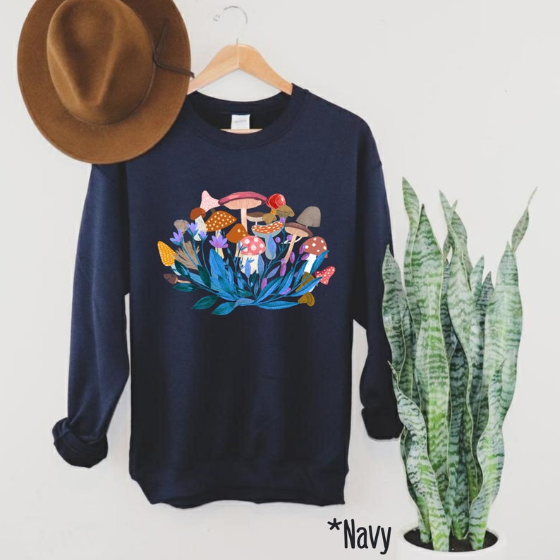 May include: Navy blue sweatshirt featuring a colourful mushroom graphic. The graphic depicts a cluster of mushrooms with red, orange, yellow, and brown caps. The mushrooms are surrounded by green leaves and purple flowers.