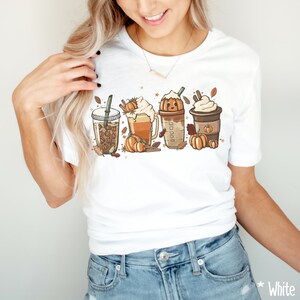 May include: White t-shirt with a graphic of four fall-themed drinks. The drinks are a pumpkin spice latte, a caramel apple cider, a pumpkin spice cold brew, and a pumpkin spice iced coffee. Each drink has a pumpkin and fall leaves around it.