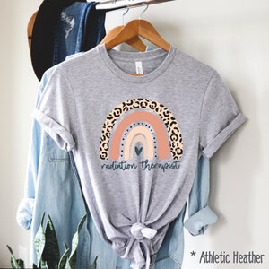 Puede incluir: Una camiseta gris con un dise&ntilde;o de arco&iacute;ris con un estampado de leopardo. El arco&iacute;ris tiene un coraz&oacute;n en el centro y el texto "radiation therapist" debajo. La camiseta est&aacute; etiquetada como "Athletic Heather".