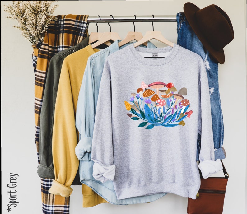 May include: A grey sweatshirt featuring a colourful mushroom and floral design. The sweatshirt has long sleeves and a crew neck.