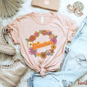 K&ouml;nnte beinhalten: Ein hellrosa T-Shirt mit einem Blumengirlanden-Design und dem Wort "Teacher" in einer Retro-Schrift.