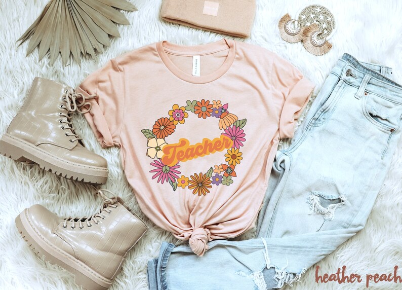 K&ouml;nnte beinhalten: Ein hellrosa T-Shirt mit einem Blumengirlanden-Design und dem Wort "Teacher" in einer Retro-Schrift.
