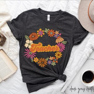 K&ouml;nnte beinhalten: Ein dunkelgraues T-Shirt mit einem floralen Kranz-Design und dem Wort "Teacher" in einer Retro-Schrift.