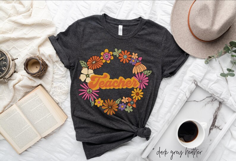 K&ouml;nnte beinhalten: Ein dunkelgraues T-Shirt mit einem floralen Kranz-Design und dem Wort "Teacher" in einer Retro-Schrift.