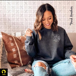 May include: A dark heather grey sweatshirt with a white outline of a monstera leaf on the front. The text "#DarkHeather" is visible in the background.
