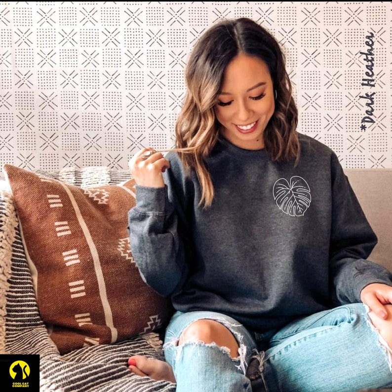 May include: A dark heather grey sweatshirt with a white outline of a monstera leaf on the front. The text "#DarkHeather" is visible in the background.