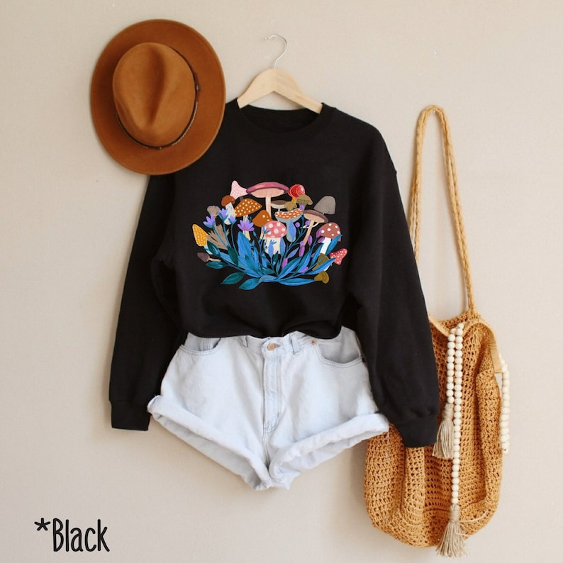 May include: Black sweatshirt featuring a colourful mushroom graphic print. The sweatshirt has long sleeves and a crew neck. The model is wearing the sweatshirt with denim shorts and a brown hat.