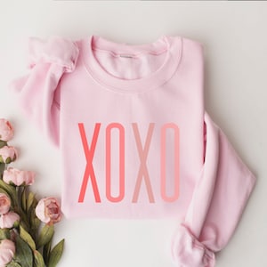 Può includere: Una felpa rosa con un design XOXO sfumato. La felpa ha un collo a volant e maniche lunghe.