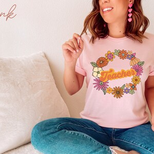 K&ouml;nnte beinhalten: Ein pinkfarbenes T-Shirt mit einem Blumengirlanden-Design und dem Wort "Teacher" in einer Retro-Schrift.