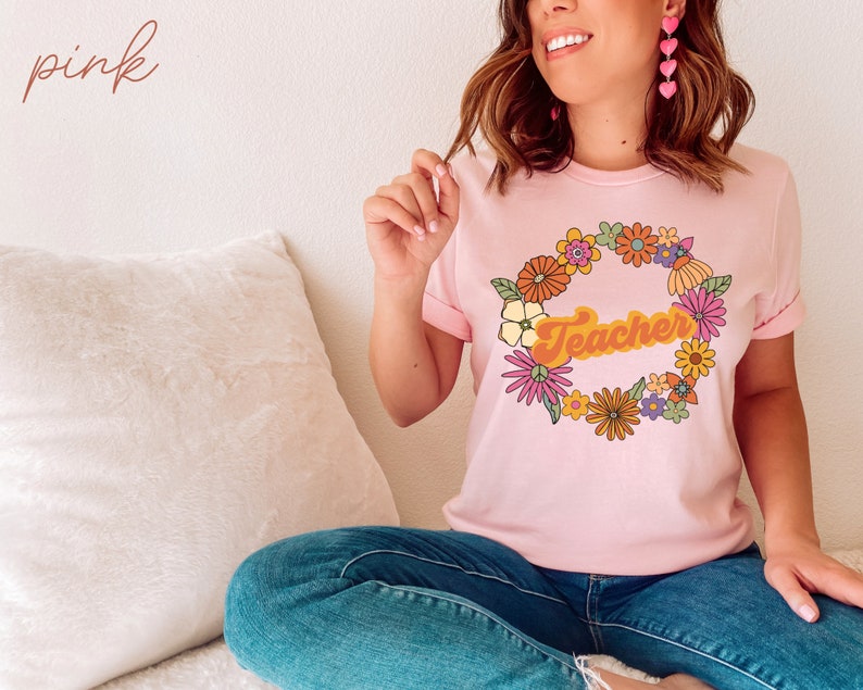 K&ouml;nnte beinhalten: Ein pinkfarbenes T-Shirt mit einem Blumengirlanden-Design und dem Wort "Teacher" in einer Retro-Schrift.