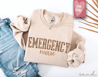 May include: A beige sweatshirt with the text "EMERGENCY nurse" in brown letters with a leopard print pattern. The sweatshirt is laid out on a white surface with a pair of blue jeans.