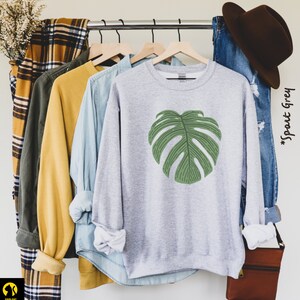 Peut inclure: Un sweat-shirt gris chin&eacute; avec un grand imprim&eacute; de feuille de monstera verte. Le texte "#Sport Grey" est visible en arri&egrave;re-plan.