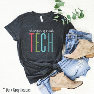 Puede incluir: Camiseta de color gris oscuro jaspeado con el texto "emergency room TECH" en letras de colores. La camiseta est&aacute; doblada y se muestra sobre una superficie blanca con un par de vaqueros azules y botines marrones.