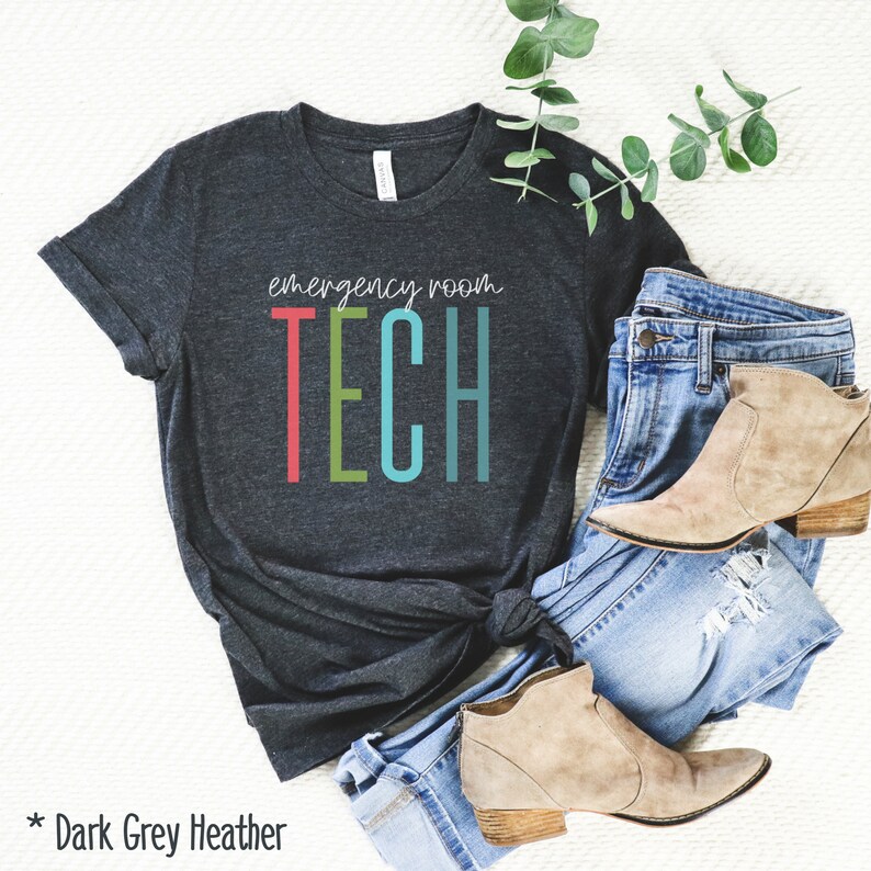 Puede incluir: Camiseta de color gris oscuro jaspeado con el texto "emergency room TECH" en letras de colores. La camiseta est&aacute; doblada y se muestra sobre una superficie blanca con un par de vaqueros azules y botines marrones.