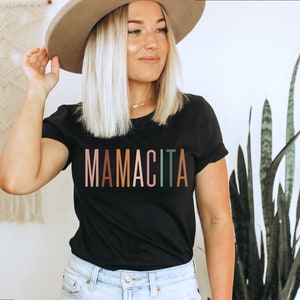 May include: A black t-shirt with the word "MAMACITA" printed in rainbow colors. The shirt is being worn by a person with blonde hair and a brown hat.