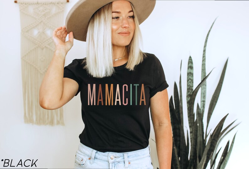 May include: A black t-shirt with the word "MAMACITA" printed in rainbow colors. The shirt is being worn by a person with blonde hair and a brown hat.