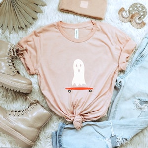 Peut inclure: T-shirt rose clair avec un motif de fant&ocirc;me blanc sur un skateboard rouge. Le t-shirt fait partie d'une collection sur le th&egrave;me d'Halloween. On aper&ccedil;oit &eacute;galement un jean en denim d&eacute;lav&eacute;, des bottines beiges et un bonnet assorti.