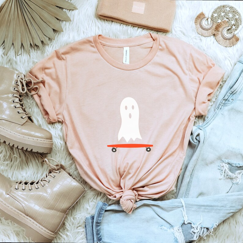 Peut inclure: T-shirt rose clair avec un motif de fant&ocirc;me blanc sur un skateboard rouge. Le t-shirt fait partie d'une collection sur le th&egrave;me d'Halloween. On aper&ccedil;oit &eacute;galement un jean en denim d&eacute;lav&eacute;, des bottines beiges et un bonnet assorti.