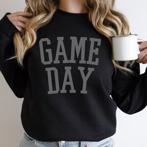 K&ouml;nnte beinhalten: Ein schwarzer Pullover mit dem Aufdruck "GAME DAY" in Grau auf der Vorderseite. Der Pullover wird von einer Person getragen, die eine wei&szlig;e Tasse h&auml;lt.