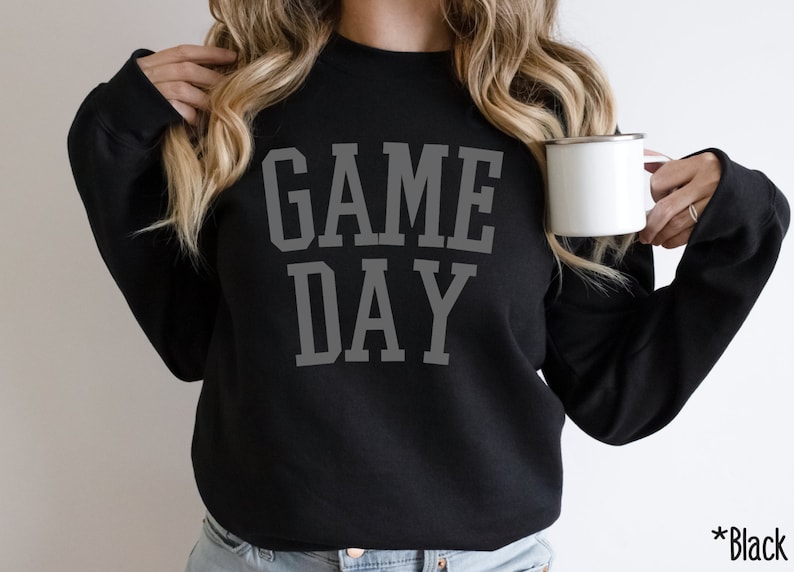 K&ouml;nnte beinhalten: Ein schwarzer Pullover mit dem Aufdruck "GAME DAY" in Grau auf der Vorderseite. Der Pullover wird von einer Person getragen, die eine wei&szlig;e Tasse h&auml;lt.