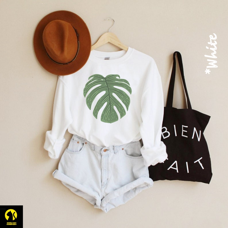 Peut inclure: Un t-shirt blanc &agrave; manches longues avec un motif de feuille de monstera verte. Le t-shirt est associ&eacute; &agrave; un short en jean bleu clair et un fedora marron. Un sac fourre-tout noir avec le texte "BIEN FAIT" en blanc est en arri&egrave;re-plan.