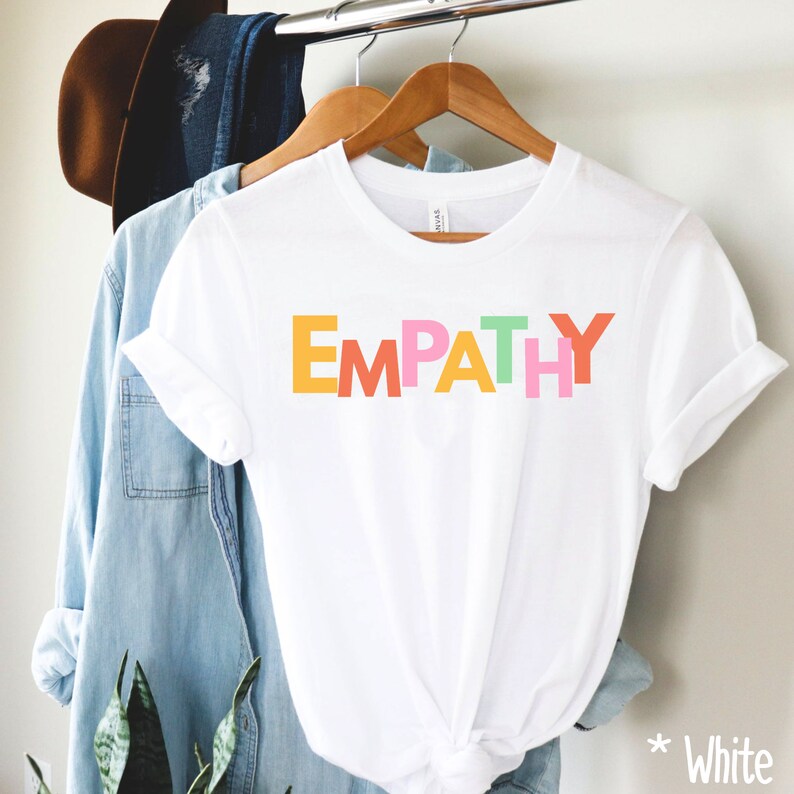May include: White t-shirt with the word "EMPATHY" printed in colorful block letters: yellow, orange, pink, and green. The shirt is hanging on a wooden hanger with other clothing items in the background.