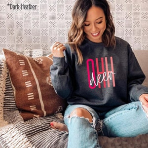 May include: A dark heather grey sweatshirt with the text "OH Deer" in pink and white lettering. The sweatshirt is being worn by a person sitting on a couch.