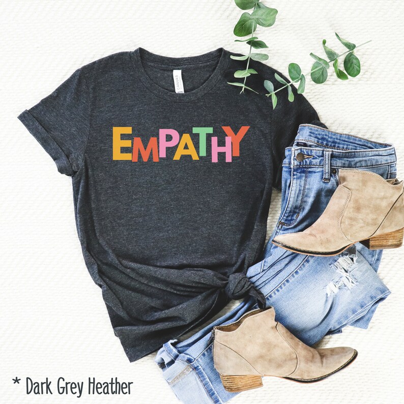 May include: Dark grey heather t-shirt with the word "EMPATHY" in colorful block letters. The shirt is styled with blue jeans and tan ankle boots. A sprig of greenery is placed on the side.