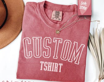 Könnte beinhalten: Ein T-Shirt in Rotbraun mit dem Text "CUSTOM TSHIRT" in Weiß. Das Shirt ist aus weichem, bequemem Stoff.
